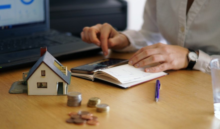 A person using a calculator beside a small model house and stacks of coins