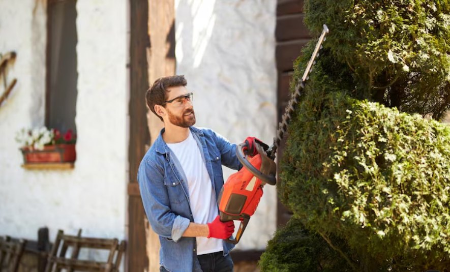 A man in safety glasses and red gloves trimming a green hedge with a power trimmer